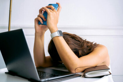 Mulher deitada sobre um computador portátil com ar desesperado e o telemóvel nas mãos para simbolizar a fadiga digital que a sociedade sofre atualmente